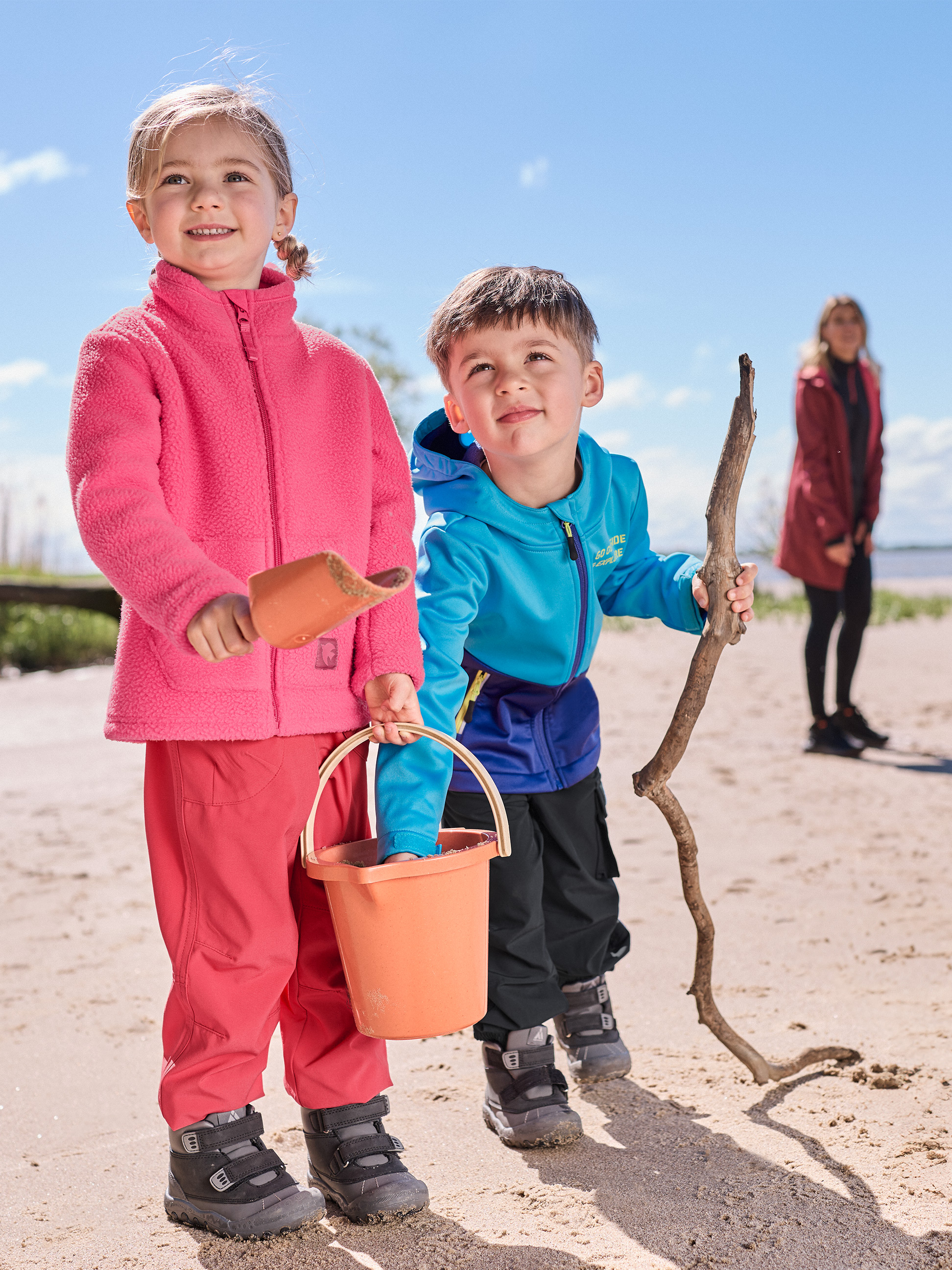 Deux enfants jouent sur la plage, vêtus de polaires roses et bleues et de pantalons imperméables.