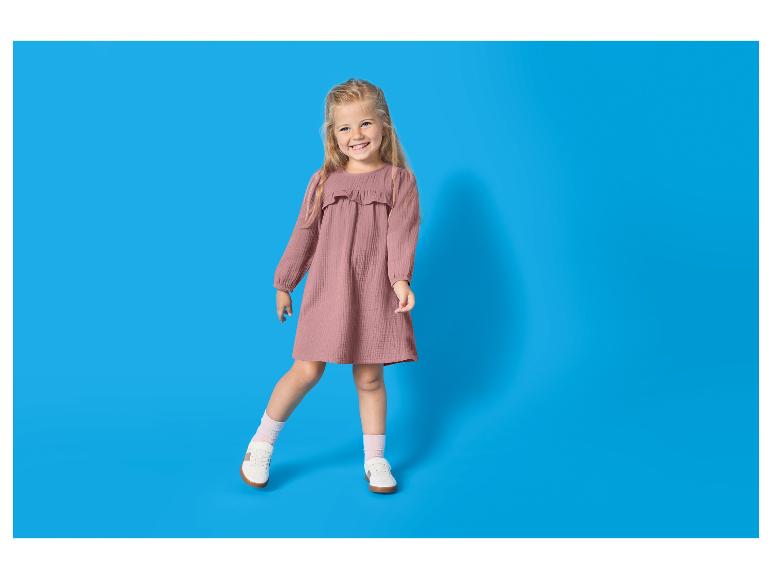 Fille souriante en robe rose et baskets blanches sur fond bleu.