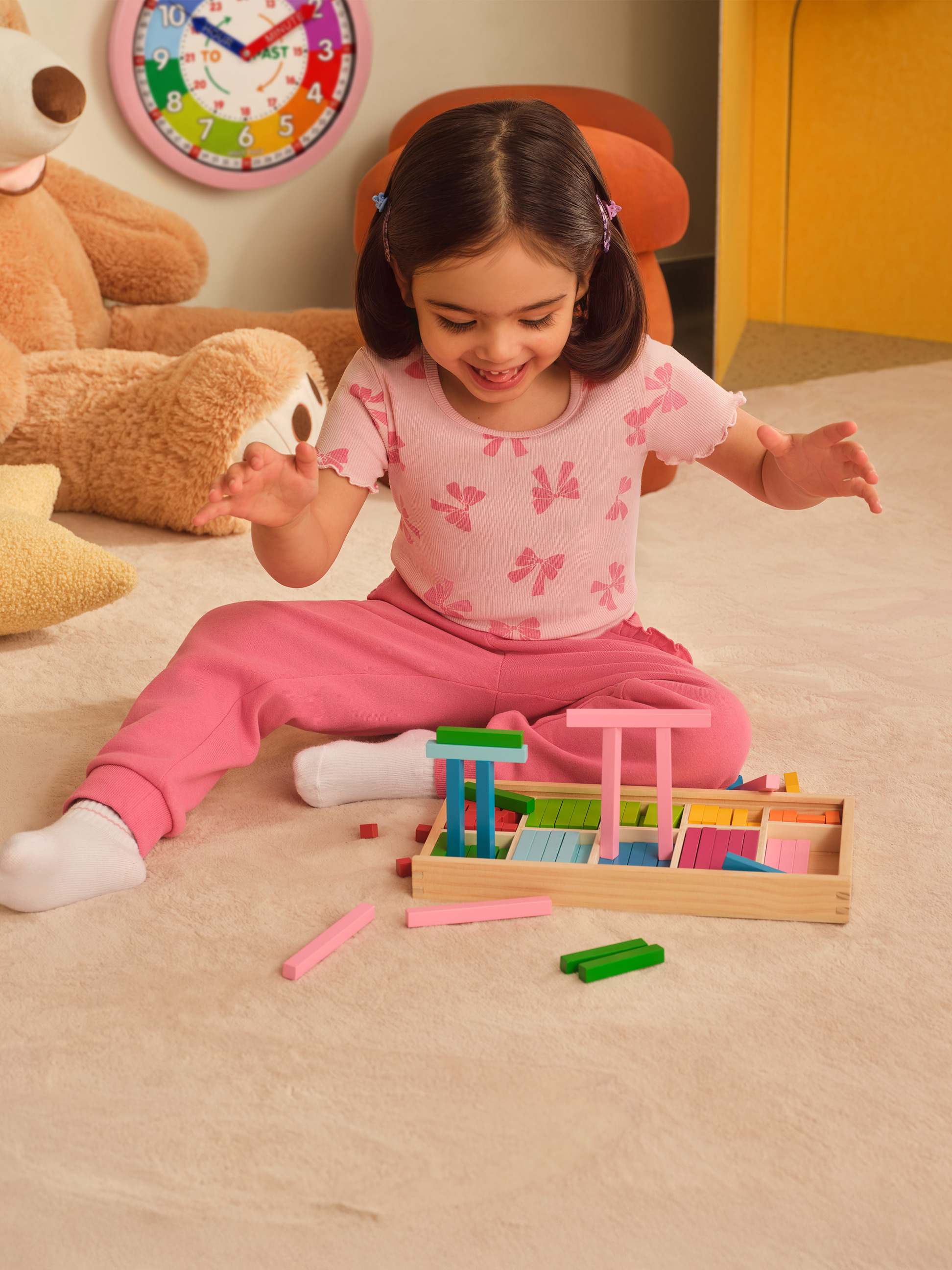 Fille souriante jouant avec des blocs de bois colorés sur un tapis, avec un ours en peluche et une horloge en arrière-plan.