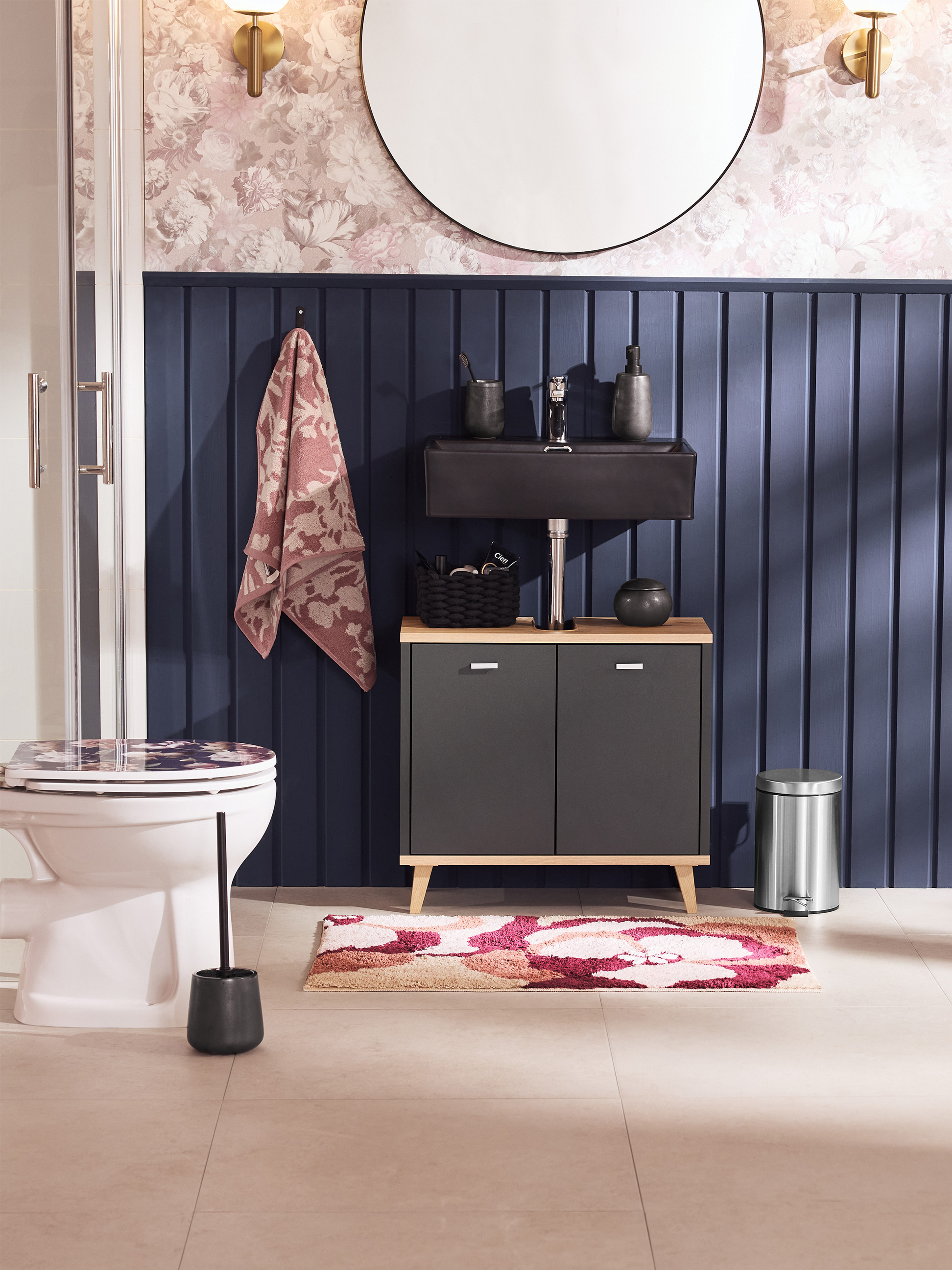 Salle de bain avec toilettes, lavabo, meuble et tapis à motifs floraux.
