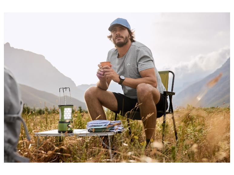 Homme en casquette et t-shirt, tenant une tasse, assis sur une chaise de camping avec une lanterne.