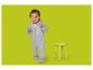 Bébé souriant en grenouillère grise, debout à côté d'un tabouret jaune.