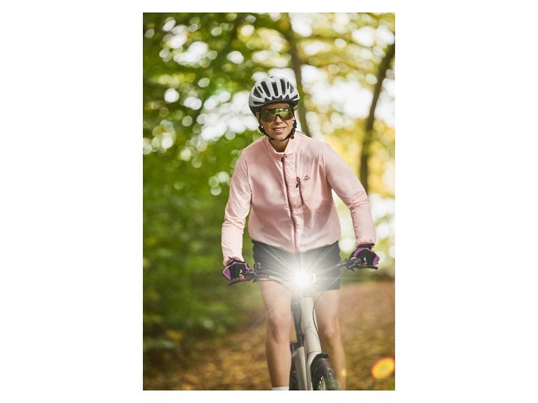 Femme souriante à vélo électrique avec casque et lunettes de soleil, vêtue d'une veste rose Crivit.
