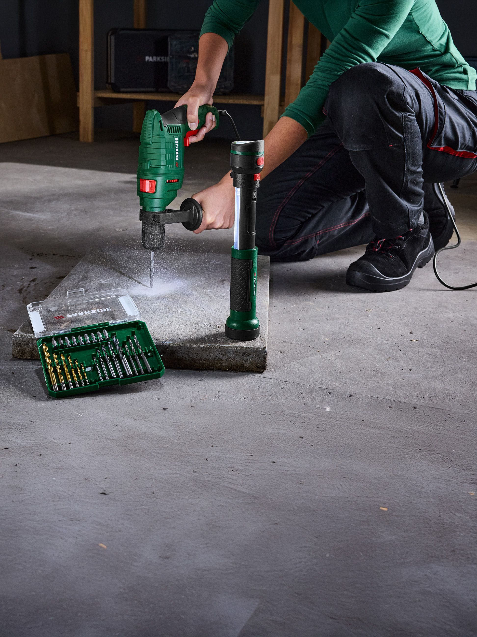 Une personne utilise une perceuse Parkside pour percer une dalle de béton, avec un jeu de forets à côté.