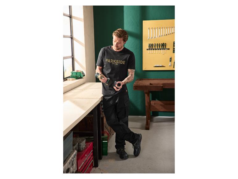Homme en t-shirt et pantalon noirs tenant une perceuse dans un atelier.