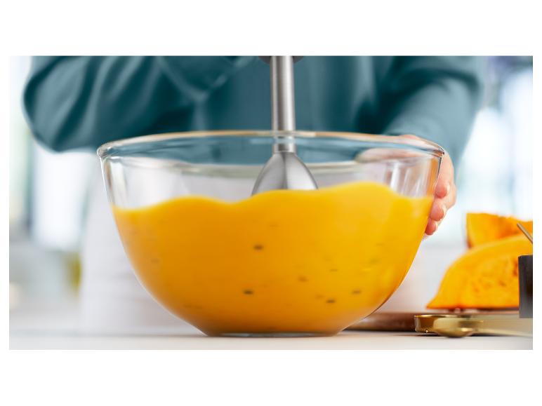 Une personne mélange une soupe à la citrouille dans un bol en verre avec un mixeur plongeant.