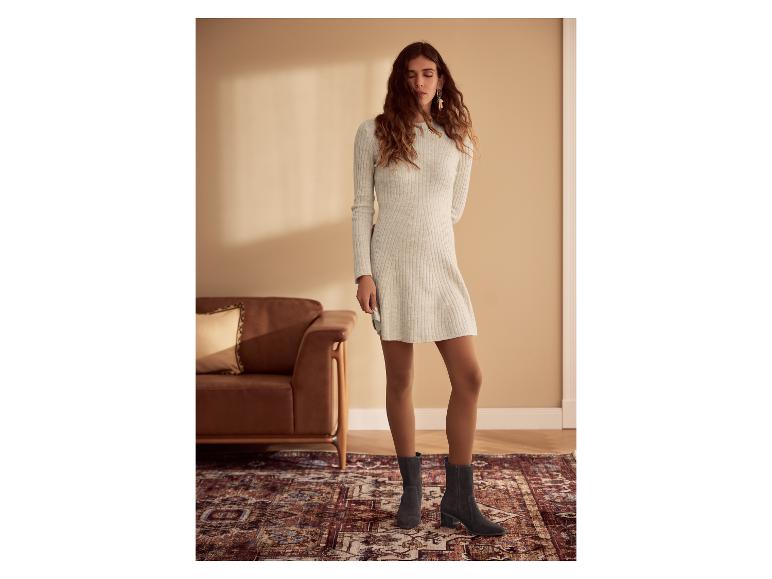 Femme en robe pull gris clair, collants marron et bottines grises, debout sur un tapis.