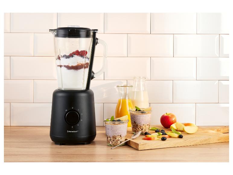 Mixeur noir avec baies et yaourt, à côté de verres avec petit-déjeuner et fruits.