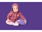 Bébé en cardigan rose à capuche et pantalon rose, assis sur un coussin violet.