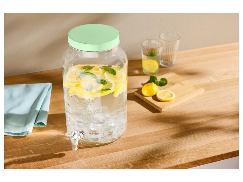 Distributeur de boissons en verre avec eau citron-menthe, verres et citrons sur une table en bois.