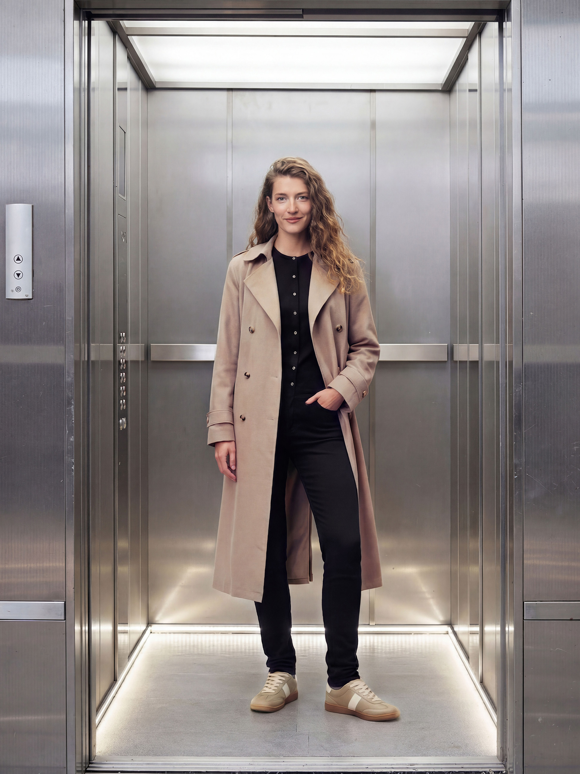 Femme en manteau beige, chemisier noir et pantalon dans un ascenseur.