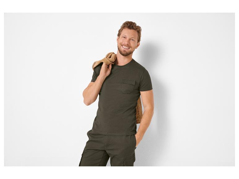 Homme souriant en T-shirt vert foncé et pantalon cargo, veste marron sur l'épaule