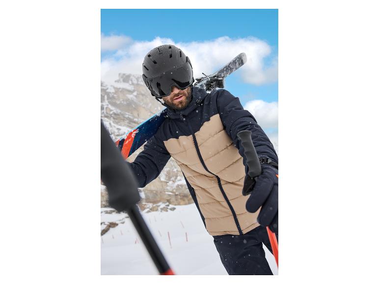 Homme avec casque et lunettes de ski, portant une veste de ski, skis sur l'épaule.