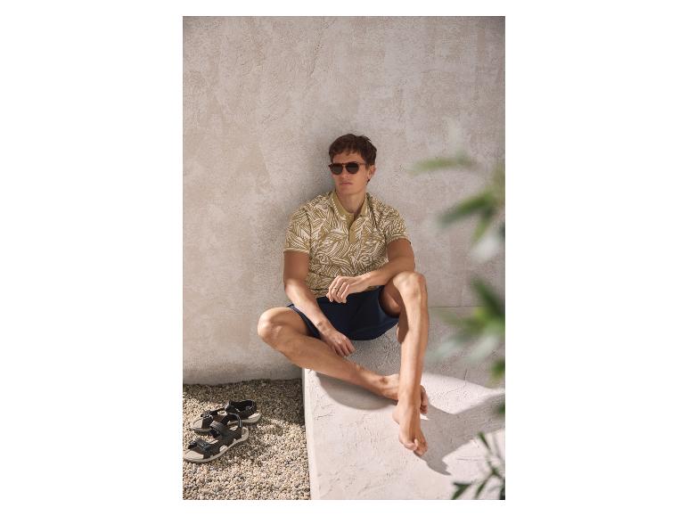 Homme en polo imprimé et short, assis avec des sandales à côté.