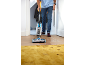 Homme nettoyant un sol en bois avec un nettoyeur multi-surfaces, se dirigeant vers un tapis jaune.