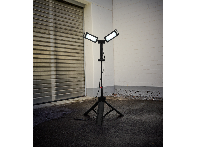 Projecteur de travail sur trépied avec deux panneaux lumineux sur l'asphalte, près d'une porte de garage et d'un mur de briques.