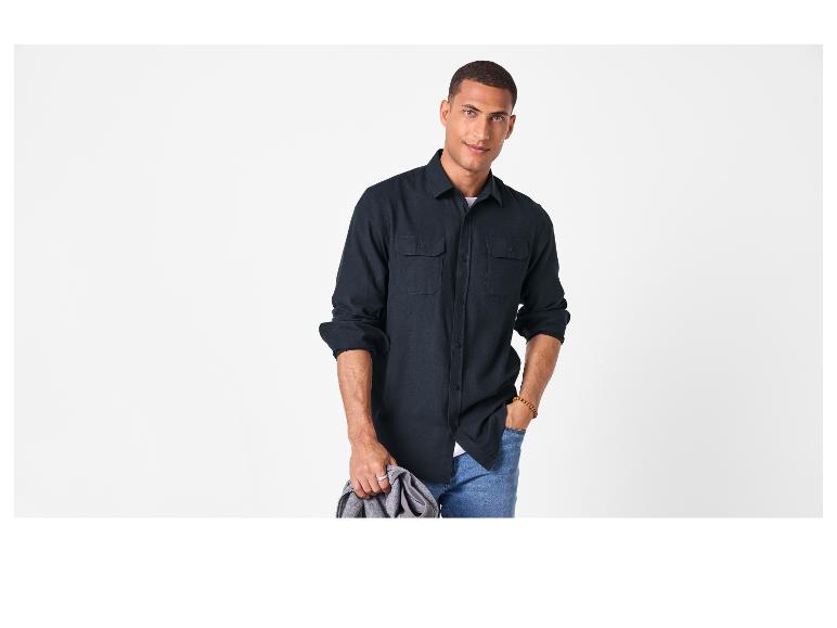 Homme en chemise foncée et jean, tenant une veste grise.