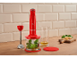 Mixeur plongeant rouge avec basilic, huile et fromage sur un comptoir en bois.