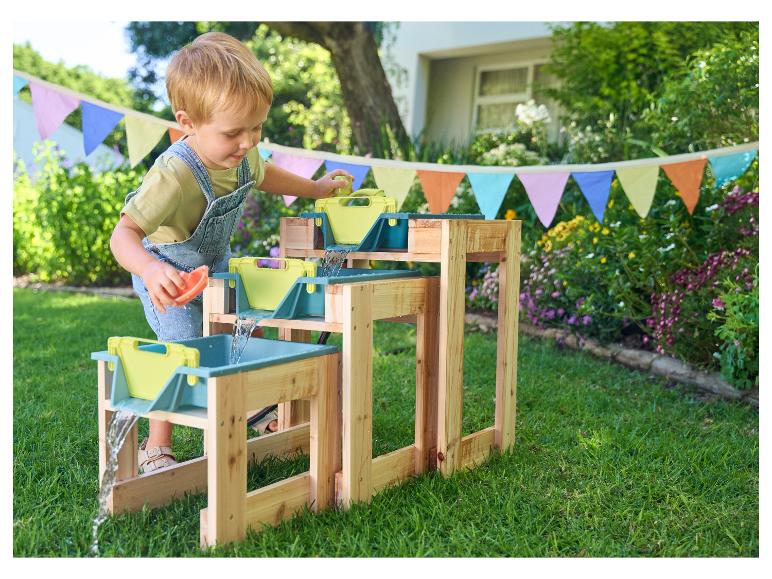 Un jeune enfant joue avec une table à eau en bois à l'extérieur, avec des drapeaux colorés en arrière-plan.