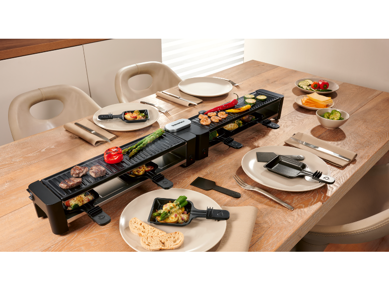 Long appareil à raclette sur une table en bois dressée avec de la nourriture et des assiettes