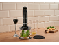 Mixeur plongeant noir avec accessoires, herbes et fromage sur un comptoir en bois.
