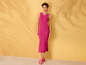 Une femme en robe rose et sandales roses pose devant un mur jaune.
