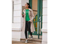 Une femme portant un trench beige, un pull vert et un pantalon noir marche dans la rue.