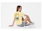 Femme en t-shirt jaune avec imprimé 'Ciao Bella' et short en jean, assise par terre.