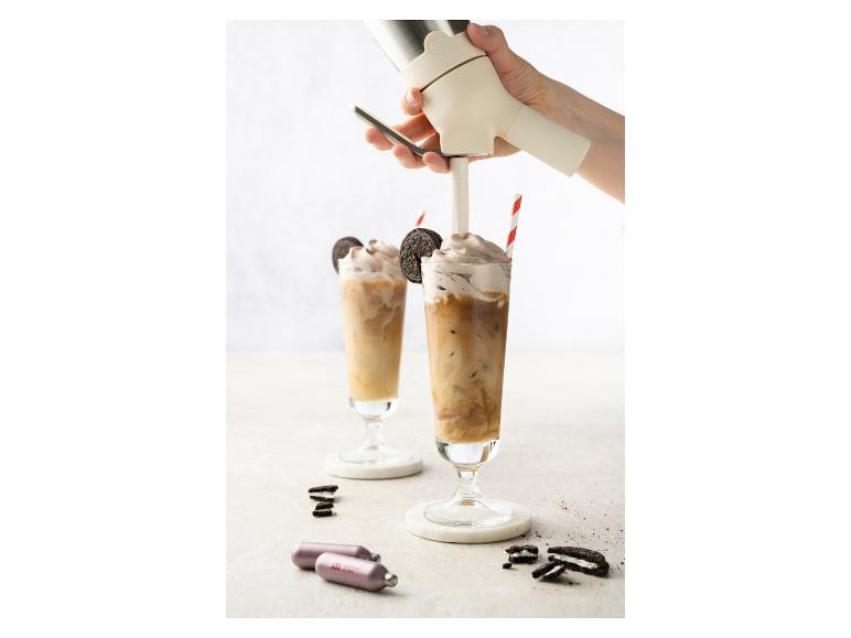 Main versant de la crème fouettée sur un café glacé avec des biscuits et des cartouches de gaz sur la table.