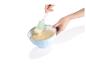 Une main prélève de la pâte à gâteau dans un bol bleu avec une spatule verte.