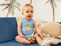 Un bébé assis sur un canapé bleu, porte un body bleu avec un soleil et joue avec des perles en bois.