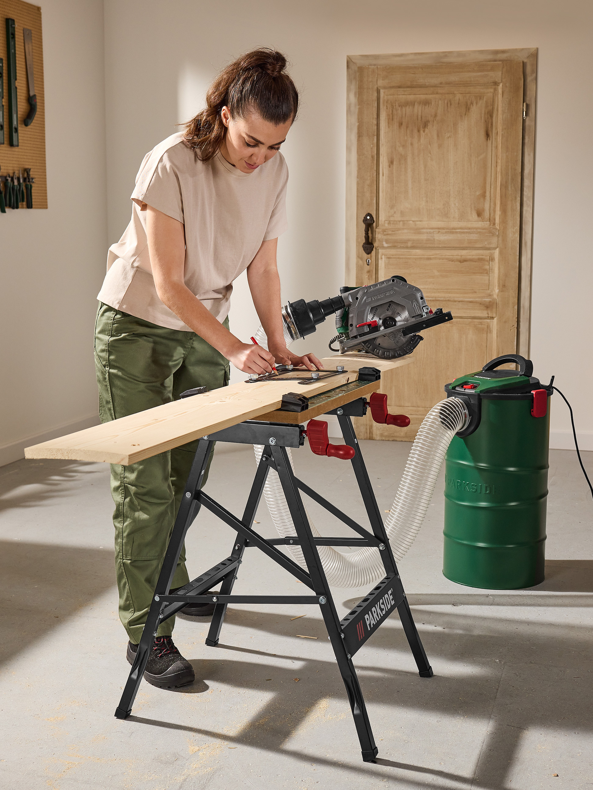 Femme marquant une planche sur un établi, avec une scie circulaire et un aspirateur industriel à côté.