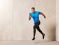 Homme en t-shirt bleu et leggings noirs courant devant un mur beige.