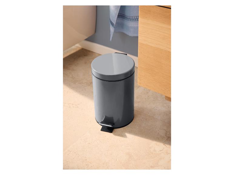 Poubelle à pédale grise dans une salle de bain avec carrelage clair et meuble en bois.