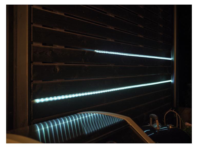 Éclairage à bande LED sur une clôture en bois sombre, se reflétant sur le couvercle d'un barbecue.
