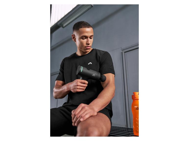 Homme en tenue de sport noire utilisant un pistolet de massage sur son bras, avec une bouteille d'eau orange à proximité.