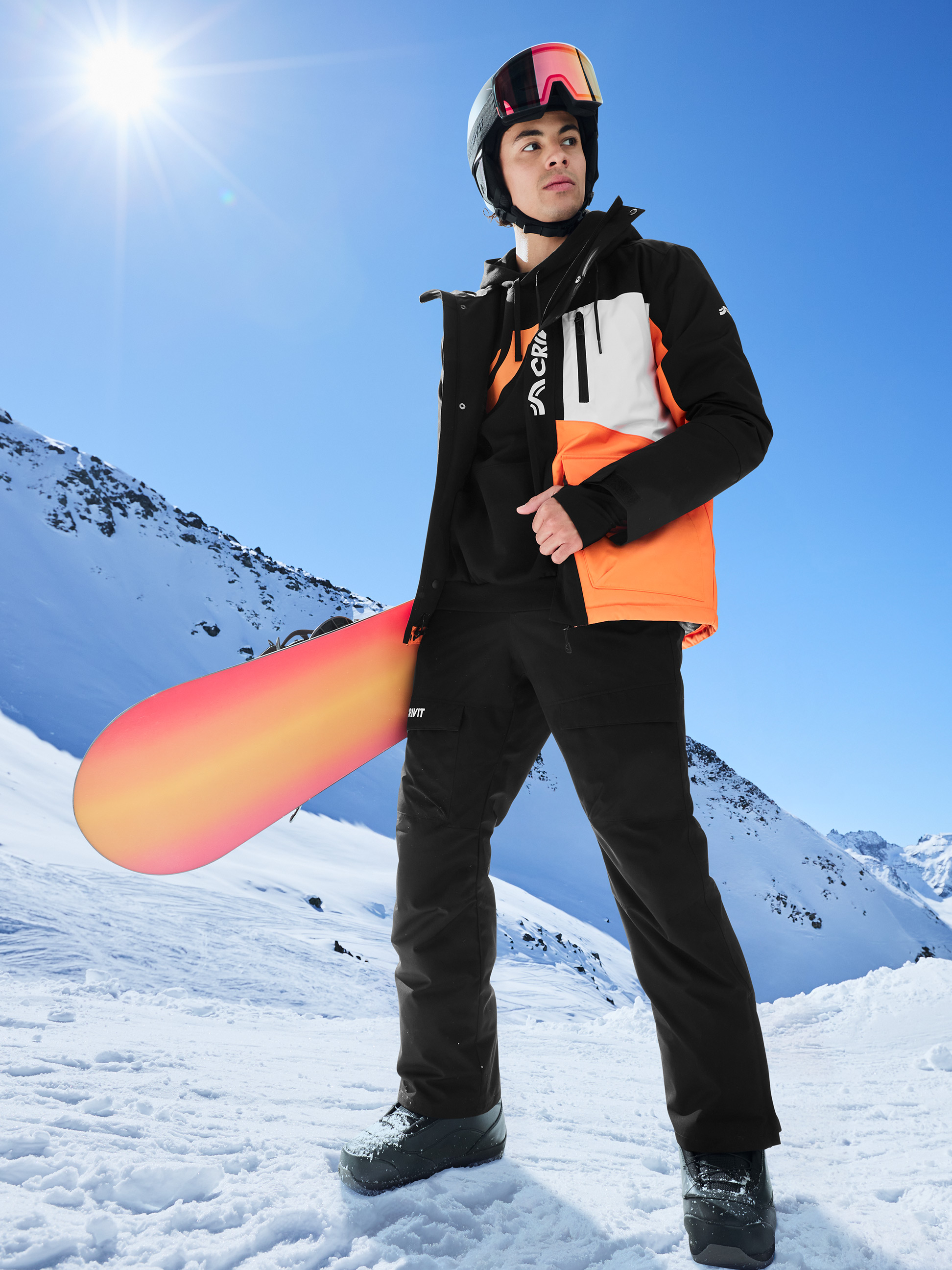 Homme en veste de ski noire et orange, pantalon de ski noir et casque avec snowboard en montagne enneigée.