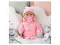 Poupée bébé en tenue rose avec sucette et biberon, entourée de coussins moelleux.