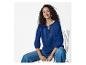 Femme en blouse brodée bleue et jean, assise sur une chaise, souriante.