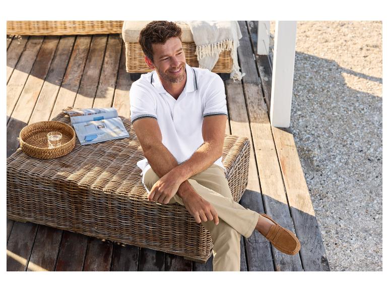 Un homme portant un polo blanc et un pantalon beige assis sur un fauteuil en rotin.