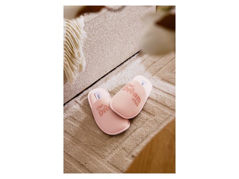 Pantoufles roses 'The Dream Club' sur un tapis beige à côté d'un canapé.