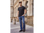 Homme en t-shirt noir et jean bleu posant devant une façade classique.