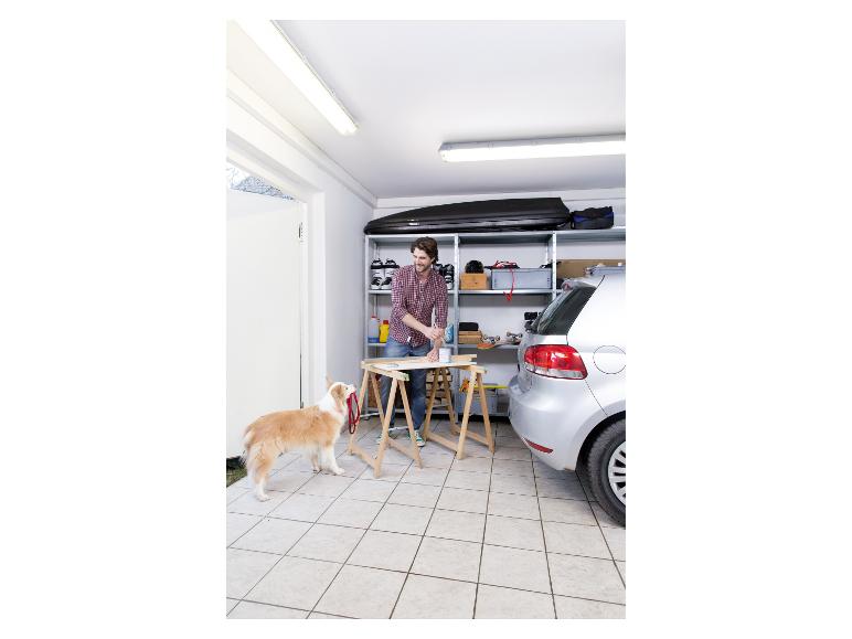Homme peignant une planche en bois dans un garage, avec un chien et une voiture à proximité.