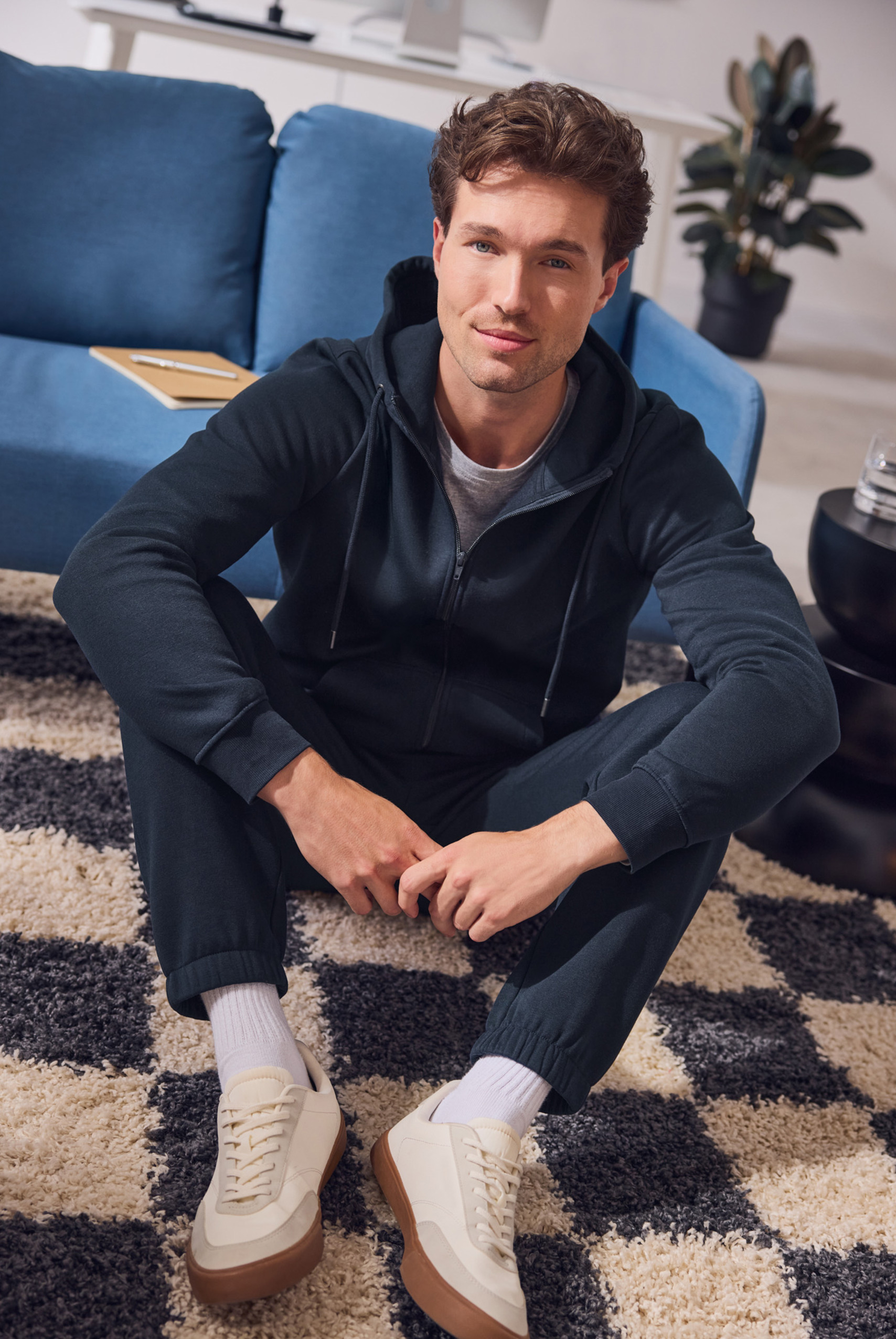 Homme en sweat à capuche et pantalon de survêtement bleu foncé assis sur un tapis à carreaux.