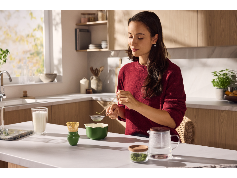 Une femme prépare du thé matcha dans la cuisine avec des ustensiles verts.