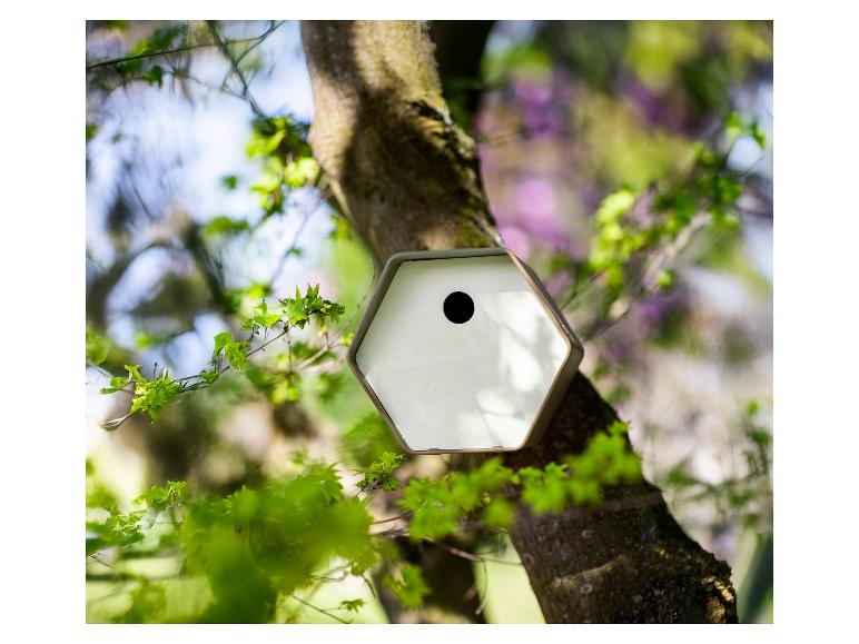 Nichoir hexagonal sur un tronc d'arbre, entouré de feuilles vertes et d'un arrière-plan flou.