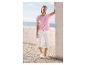 Femme en t-shirt rose perforé et pantalon capri blanc, appuyée contre un mur avec vue sur la mer.