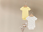 Deux bodies bébé, l'un avec un motif d'abeilles et l'autre jaune uni, suspendus à un cintre en bois.