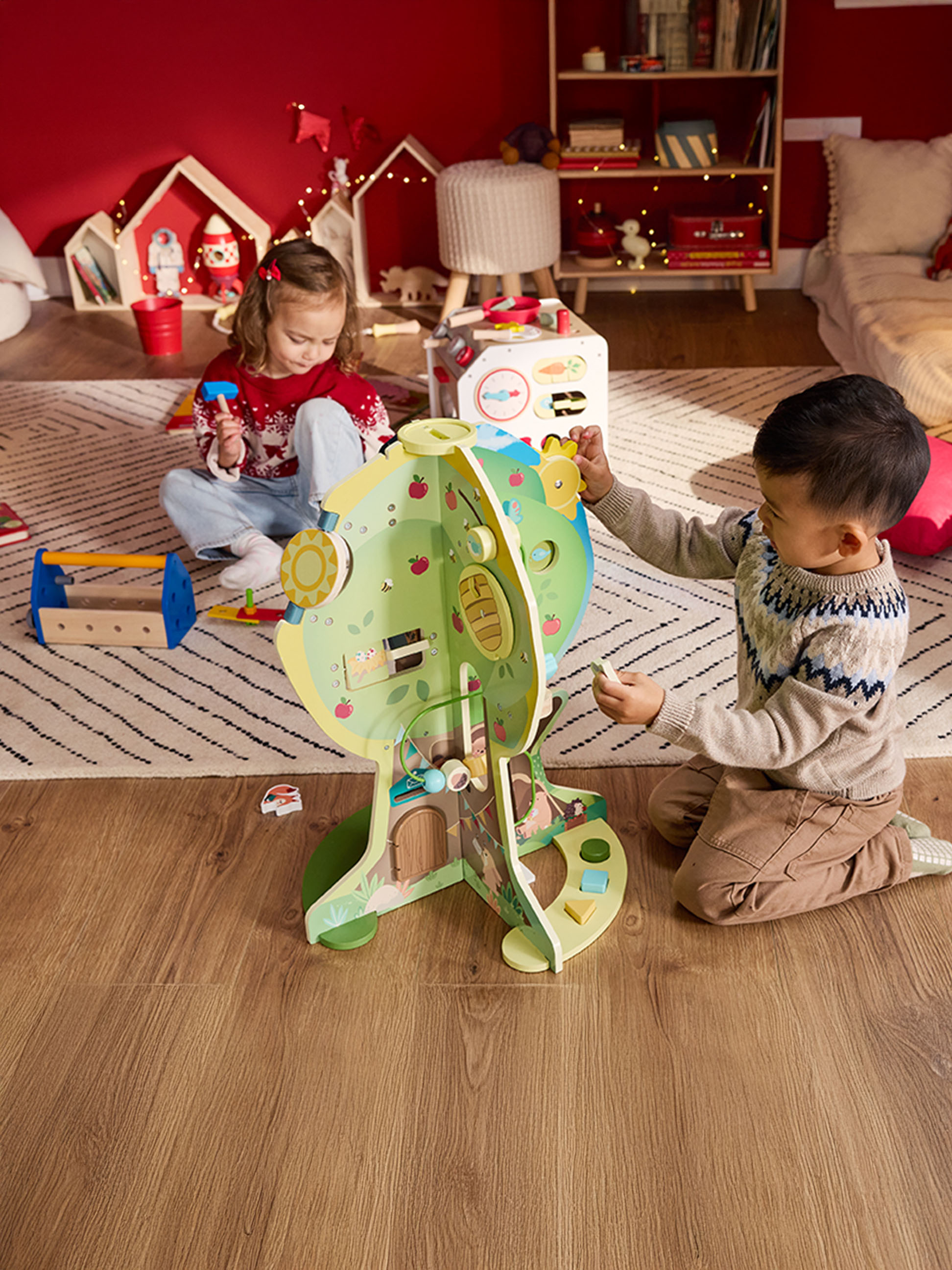Deux enfants jouent avec une maison de poupées en bois et une boîte à outils dans une pièce décorée pour les fêtes.
