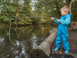 Un garçon en salopette imperméable bleue pêche à la ligne avec un bâton et une corde dans un ruisseau.